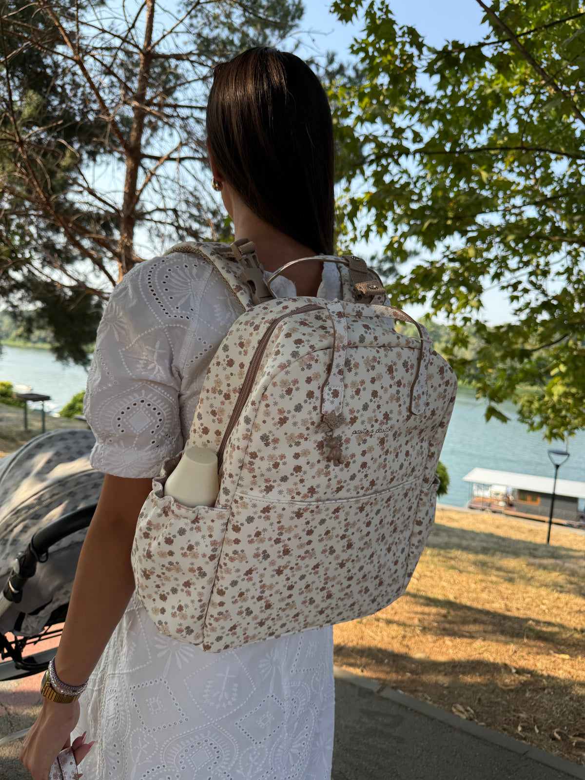 A woman viewed from behind wearing a cream-colored backpack with a brown floral pattern, standing outdoors near a lake with trees in the background.
