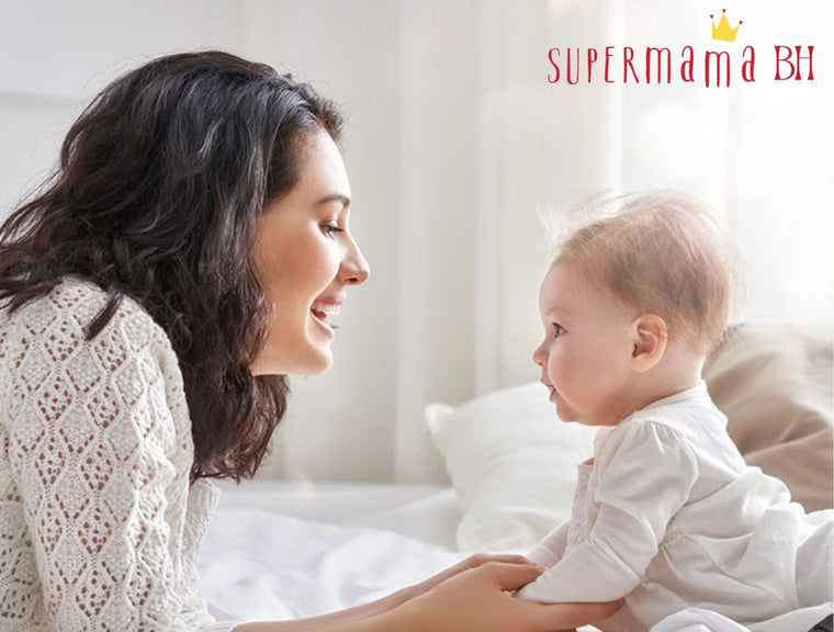 Woman and baby sitting together on a bed with 'Supermama BH' logo.